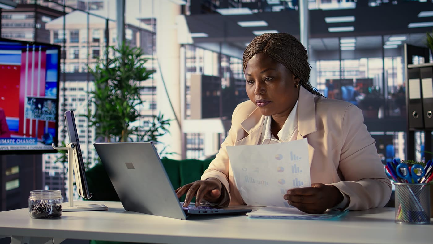 Businesswoman comparing a printed report with data on her laptop while organizing subscription information